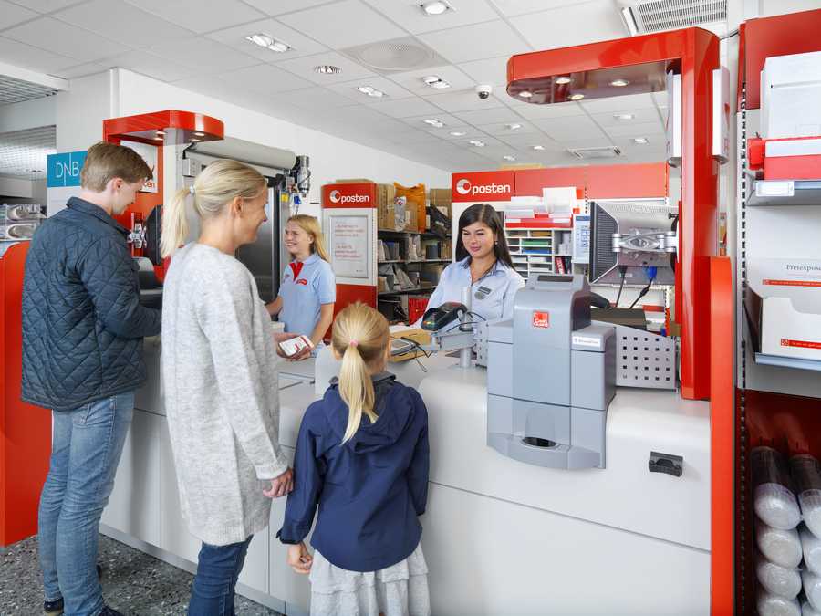 Flere kunder står på et postkontor med to butikkansatte bak skranken.